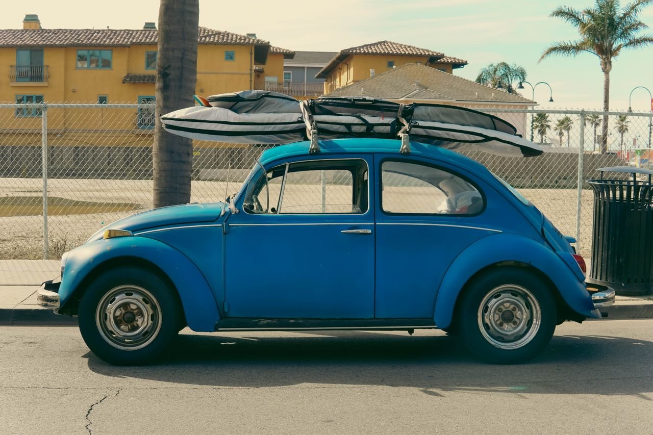 A classic blue Volkswagen Beetle with surfboards parked by a sunny beachside in California.